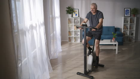 El-Hombre-De-Pelo-Gris-Está-Entrenando-En-Bicicleta-De-Spinning-En-La-Sala-De-Estar-Con-Un-Estilo-De-Vida-Saludable-Y-Manteniendo-Una-Buena-Condición-Física-A-Mediana-Edad